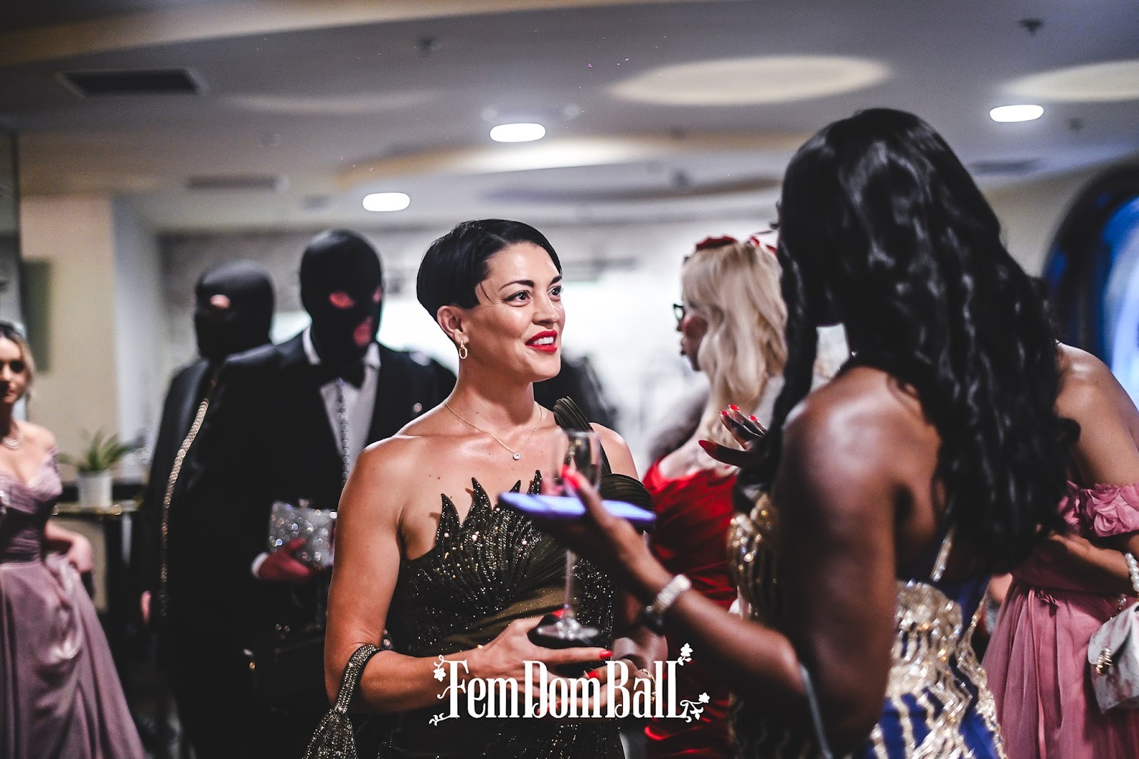 Zwei Dommes im Gespräch beim Femdom Ball, im Hintergrund servieren zwei submissive Männer Getränke